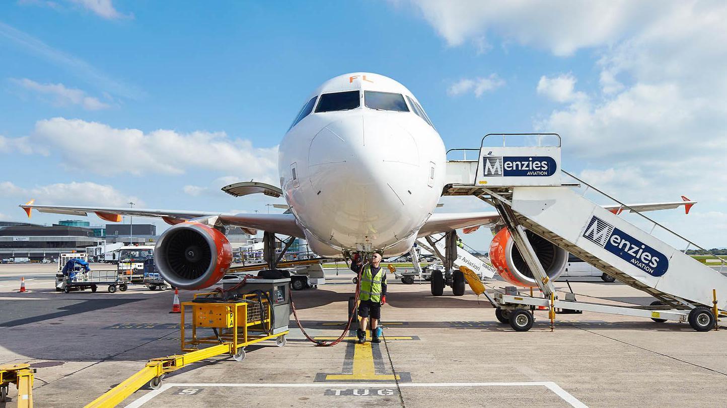 Vista frontal de um avi&atilde;o comercial estacionado, com equipes de solo trabalhando sob o nariz da aeronave e uma escada m&oacute;vel com a marca &ldquo;Menzies Aviation&rdquo; posicionada ao lado