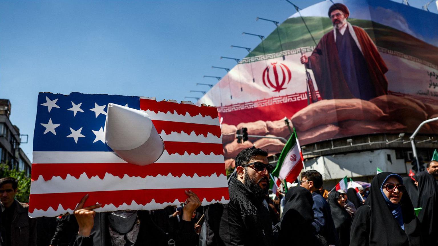 Uma pessoa segura um cartaz representando a bandeira dos EUA, com uma imagem do novo L&iacute;der Supremo do Ir&atilde;, Mojtaba Khamenei, em um outdoor ao fundo, no dia da cerim&ocirc;nia que marcou os 40 dias desde o assassinato do Aiatol&aacute; Ali Khamenei.