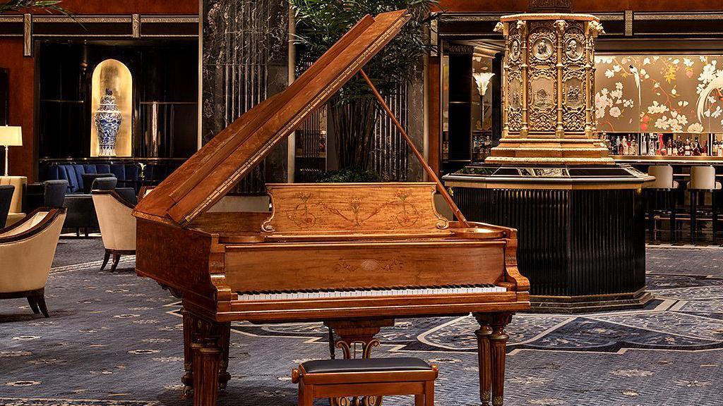 Un piano de cola de madera marrón en el centro, detrás un antiguo reloj dorado y butacas de madera con tapizado color crema.
