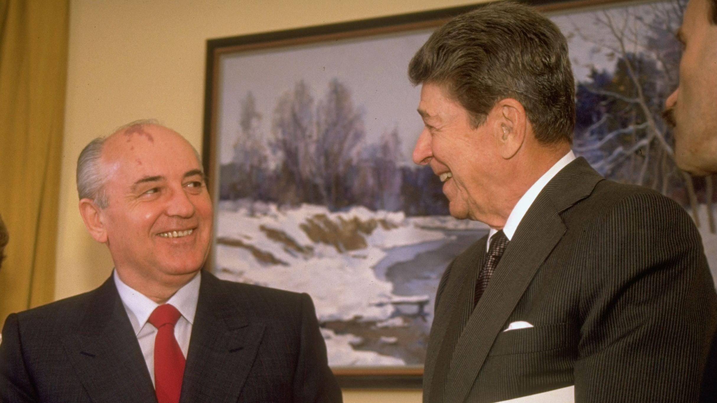 USSR President Mikhail Gorbachev, wearing a navy suit, light-blue shirt and red tie, meets US President Reagan, in a dark-grey pin-striped suit, with white pocket handkerchief, white shirt and dark, shiny, patterned tie, in San Francisco, on 4 June 1990. The two men are smiling warmly as they look at each other. A painting of a snowy landscape hangs in the background.