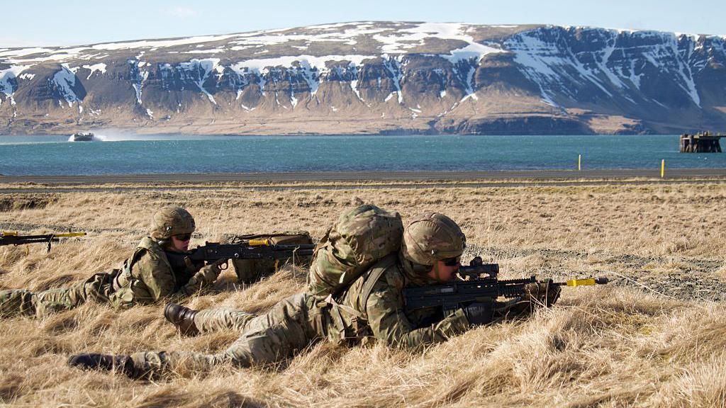 Dois soldados americanos est&atilde;o deitados, apontando suas armas em um campo. A que est&aacute; atr&aacute;s parece ser uma mulher. Ao fundo, v&ecirc;-se montanhas nevadas.