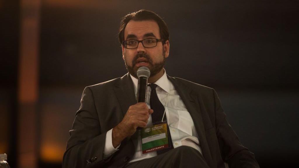 Christopher Garman sentado e falando ao microfone durante um evento.