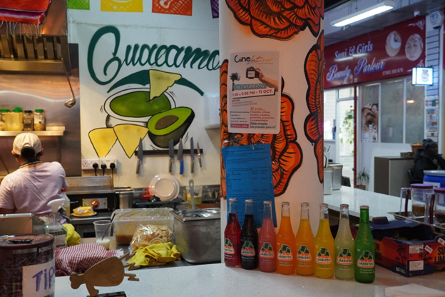 Se ve un mostrador del restaurante. y sobre este una hilera de botellas con soda. en la pared del fondo hay una ilustración de un aguacate con tortillas y la palabra "guacamole". Se a una empleada de espaldas cocinando. 