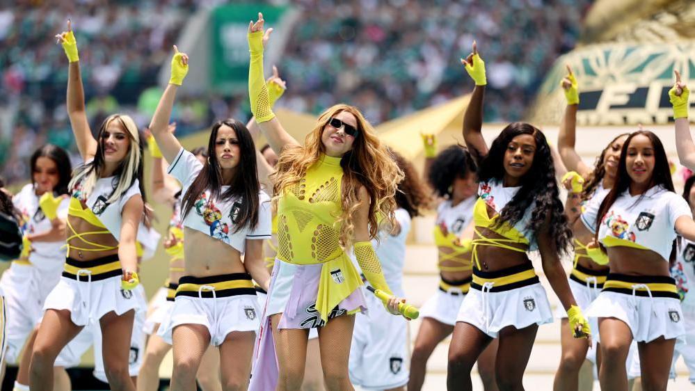 Shakira, mengenakan rok pendek putih dan atasan jaring kuning serta sarung tangan, tampil bersama sekelompok penari dengan busana serupa