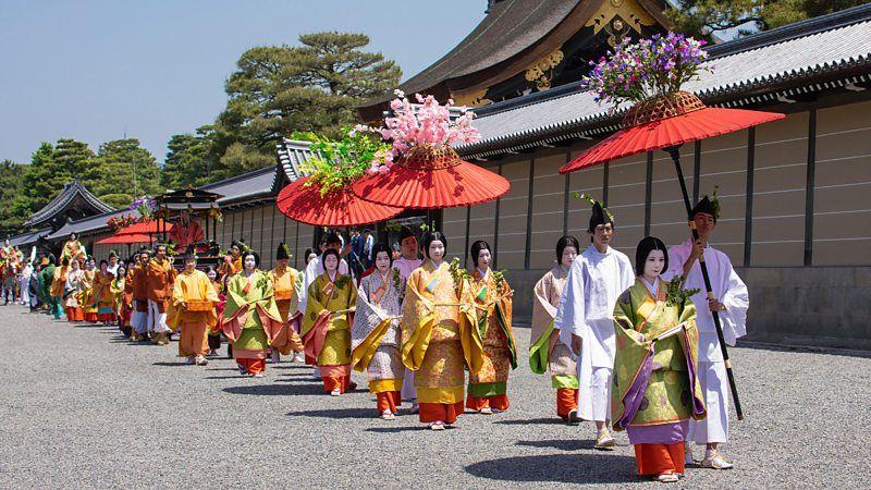 Desfile pelas ruas de cidade do Japão, com pessoas portando guarda-chuvas vermelhos de cabos longos