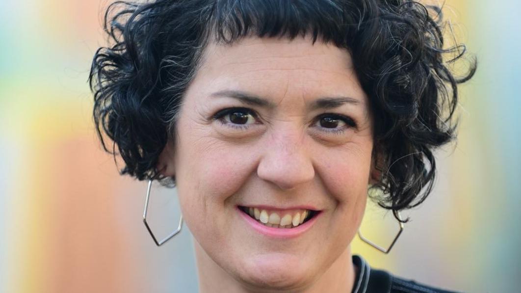 勞拉墨菲是一位年輕女子，她有著黑色捲髮和八角形銀色耳環，在淺藍色背景的襯托下對著鏡頭微笑。