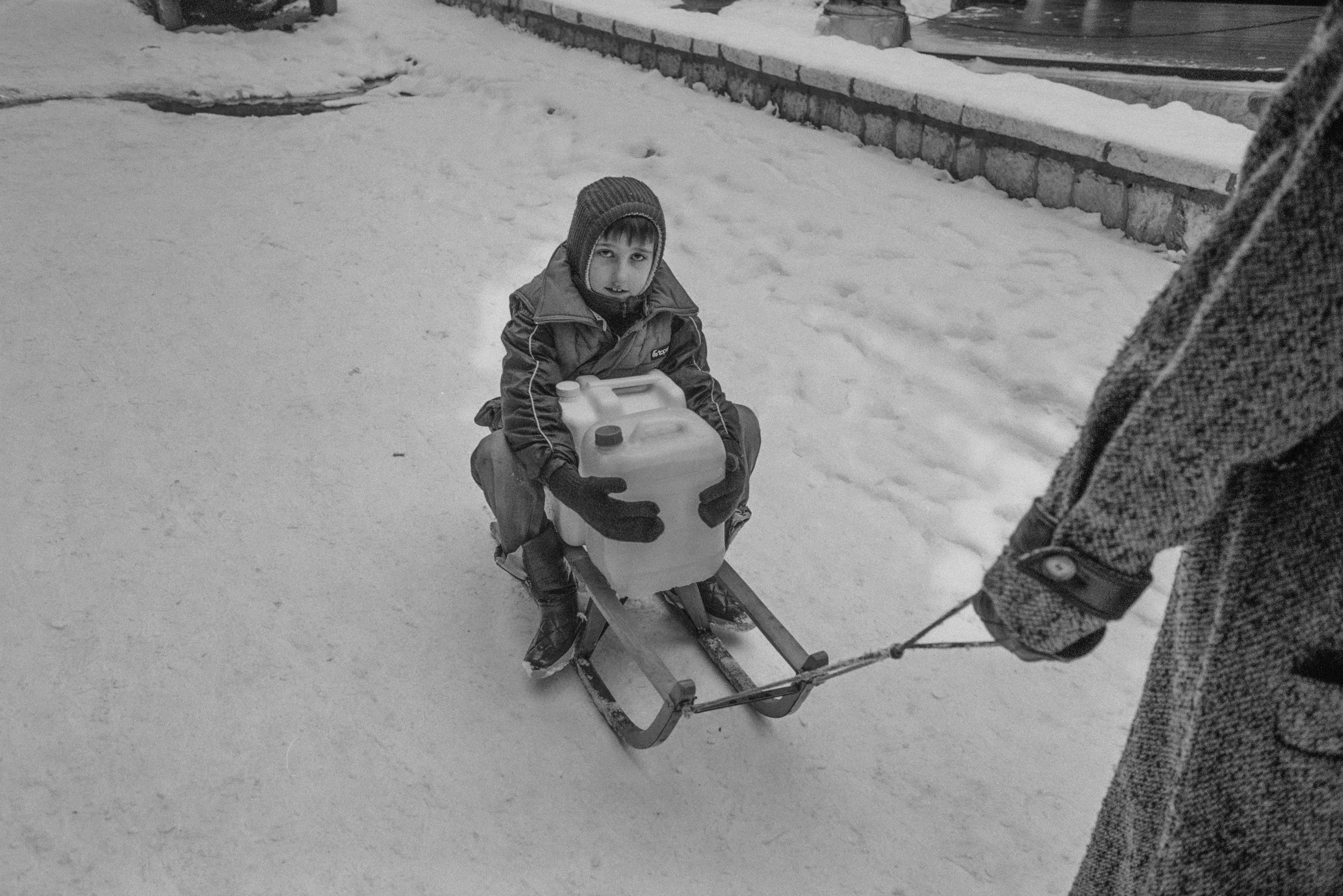 겨울에 물통을 안고 썰매를 타는 어린 아이의 흑백 사진