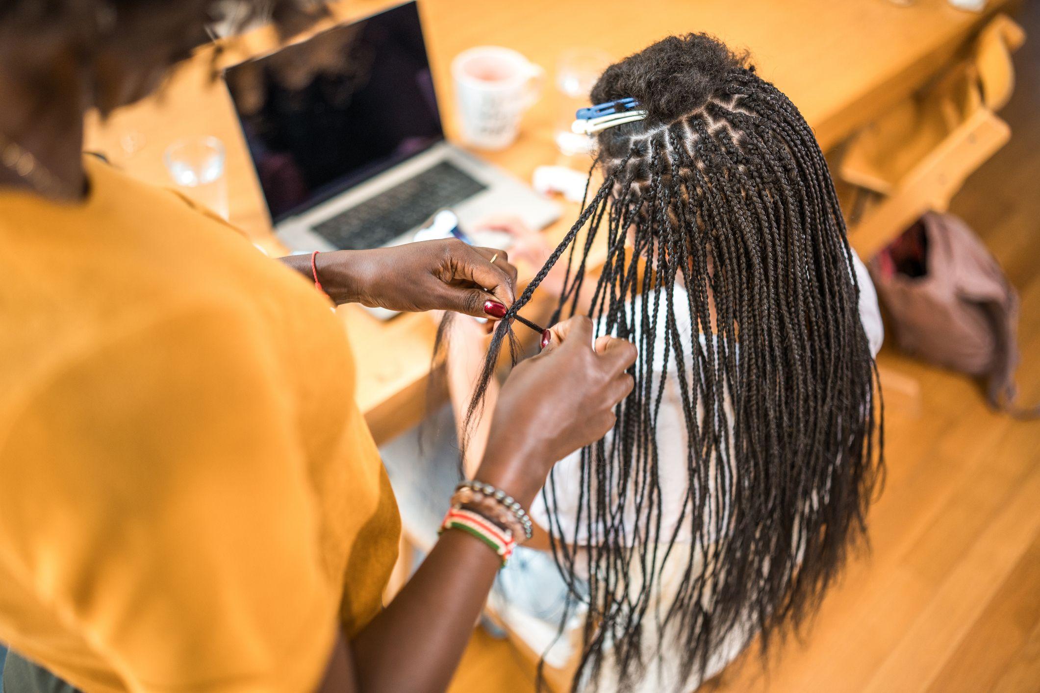 Bird eye view of a woman seated while having braids installed. The bottom part is already braided, and the stylist is working her way up to the top of the head which has natural hair clipped up. 