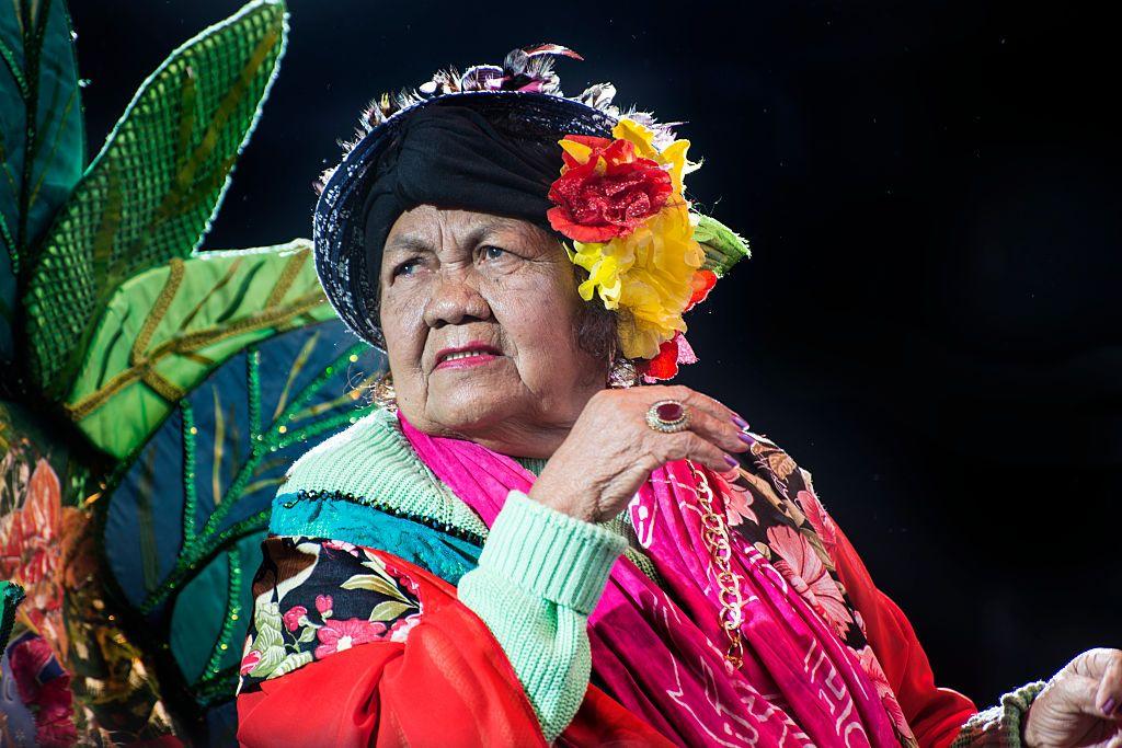 Dona Onete com roupas coloridas e anel no dedo; ao fundo, uma decora&ccedil;&atilde;o inspirada em folhas 