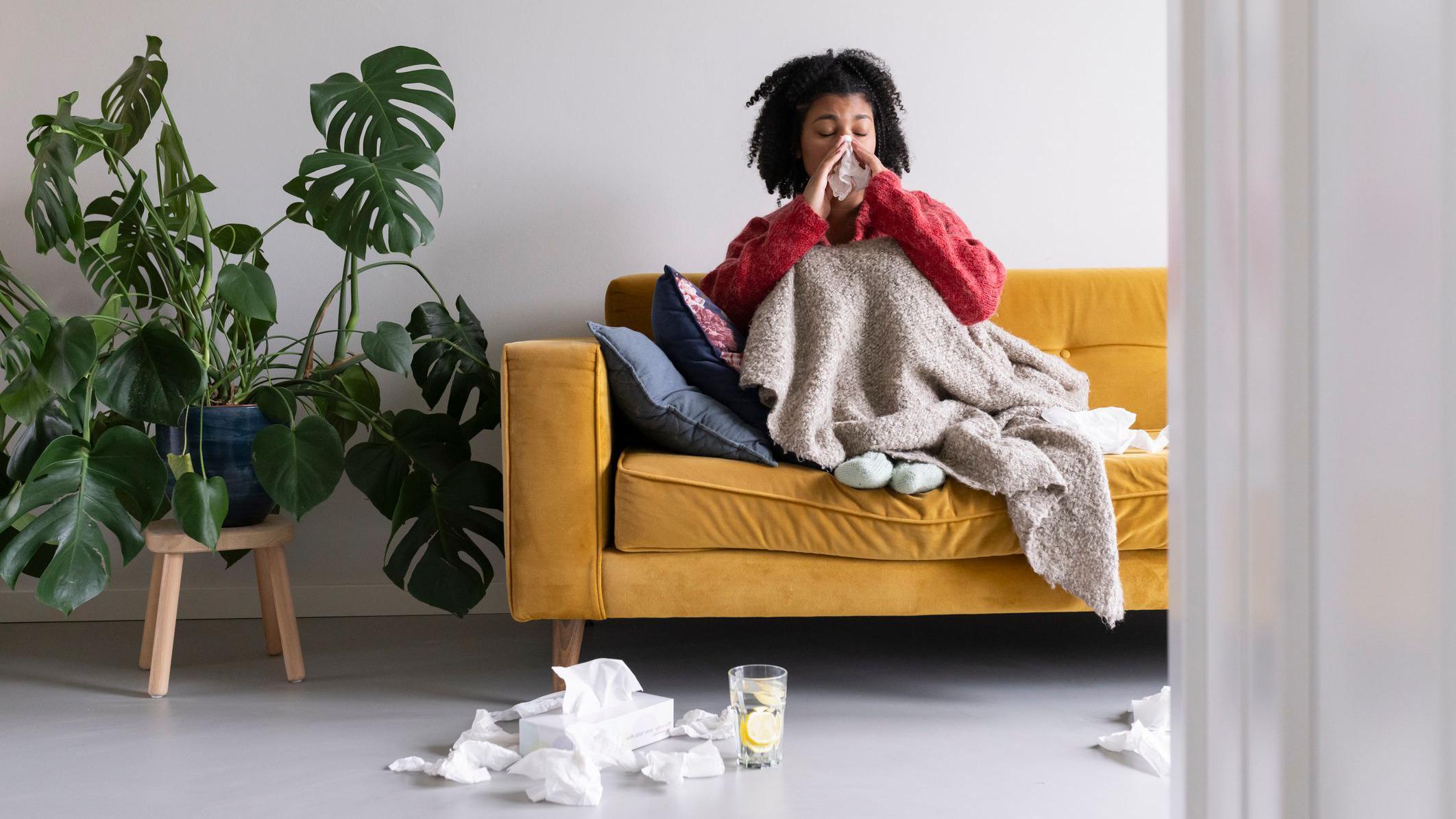 Uma pessoa sentada em um sofá amarelo, usando uma blusa vermelha, debaixo das cobertas assoa o nariz. No chão estão lenços de papel e um copo de água com uma rodela de limão.