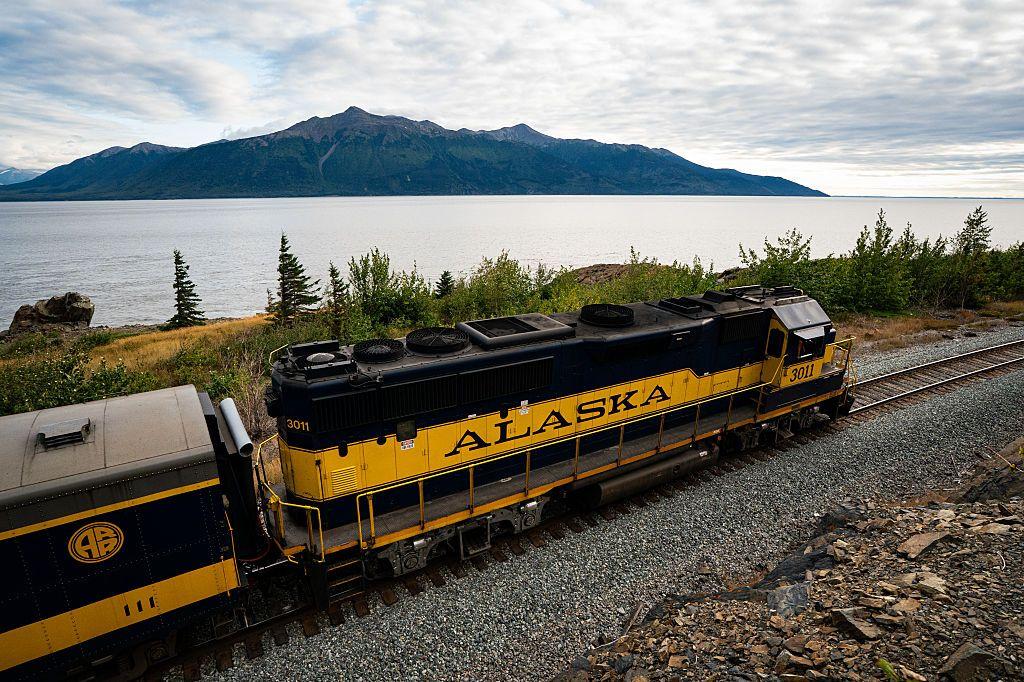Un tren negro y amarillo con la palabra Alaska escrita en la locomotora, junto a lo que parece ser el mar. Al otro lado se divisa una montaña. 