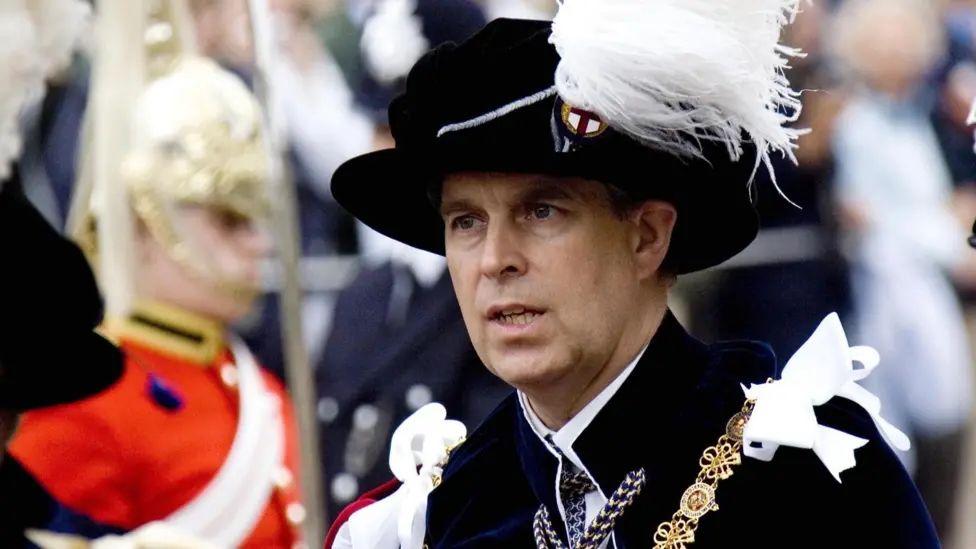 Andrew wearing finery in front of a royal guard. Andrew has a black hat with a white feathery design. He has a gold sash over his left shoulder and a black jacket. The royal guard is standing to attention, wearing a red tunic with a white sash across his chest. He has a helmet with white feather-like material on the top and falling down the side of his head.