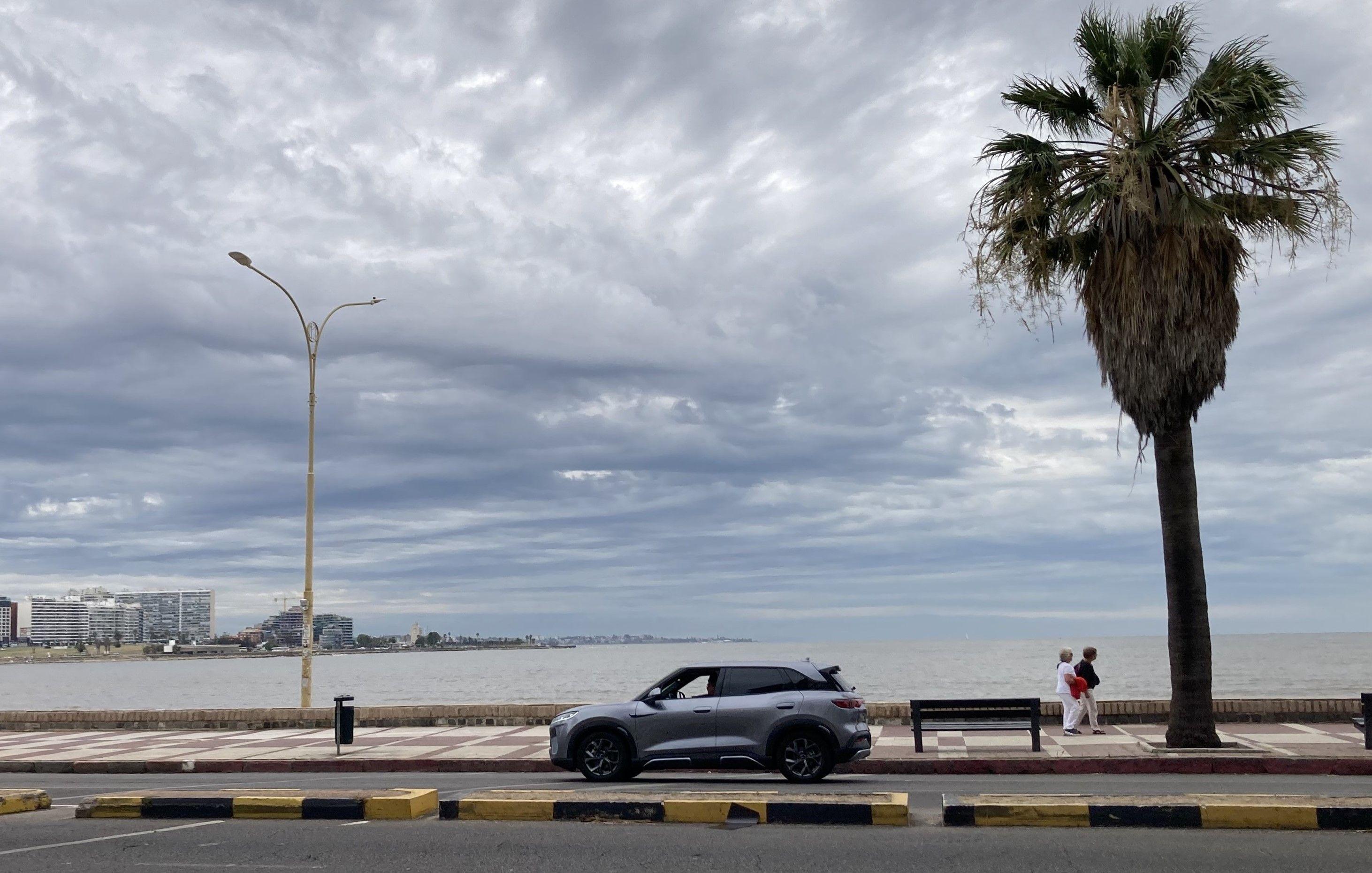 Un auto eléctrico pasa una palmera y dos personas por la rambla de Montevideo.