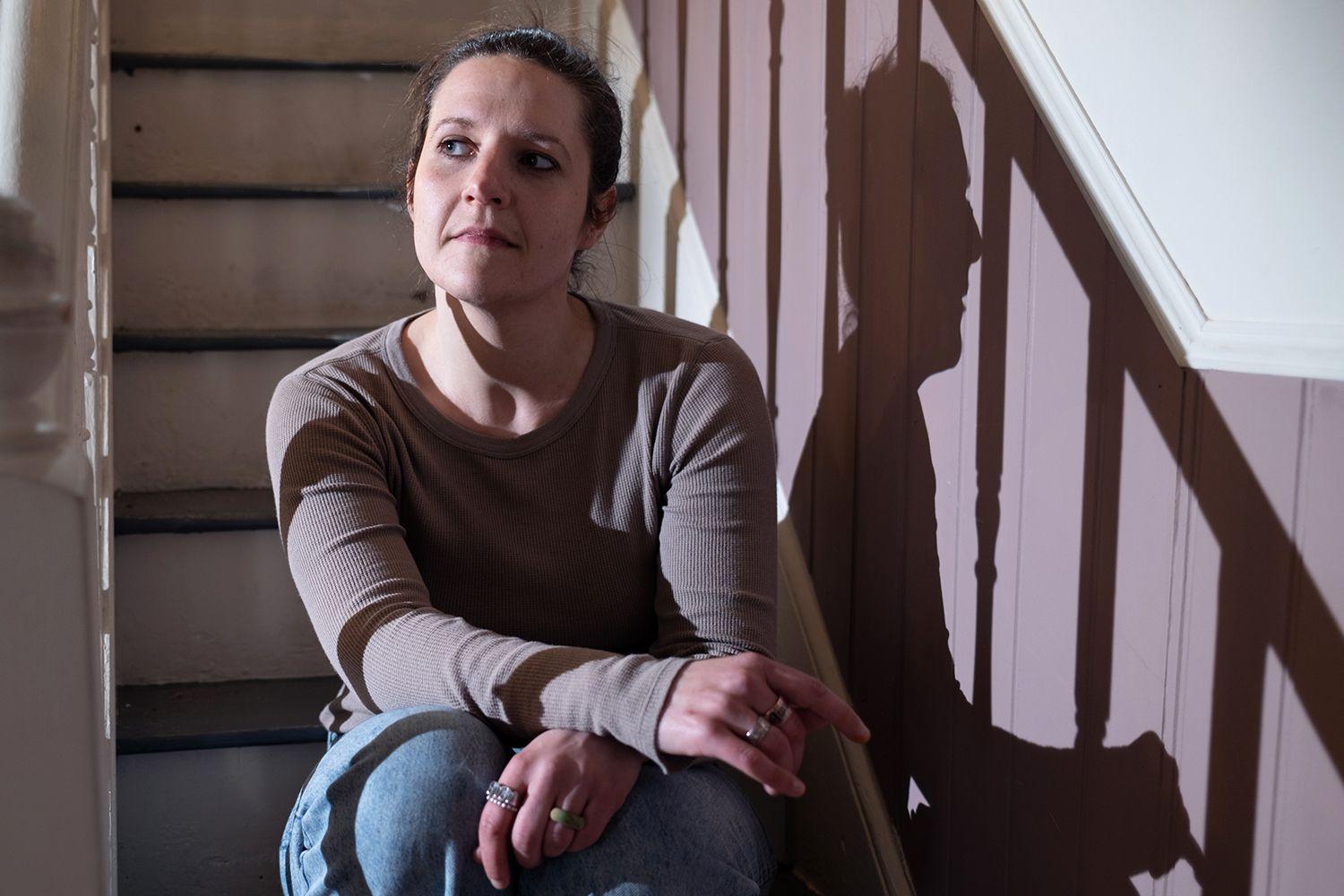 Ruth est&aacute; sentada na escada de uma casa. Iluminada por uma luz lateral, que projeta sua sombra sobre uma parede com pain&eacute;is, ela olha levemente para o lado, com um dos bra&ccedil;os apoiado sobre o joelho e veste uma blusa de mangas compridas e jeans