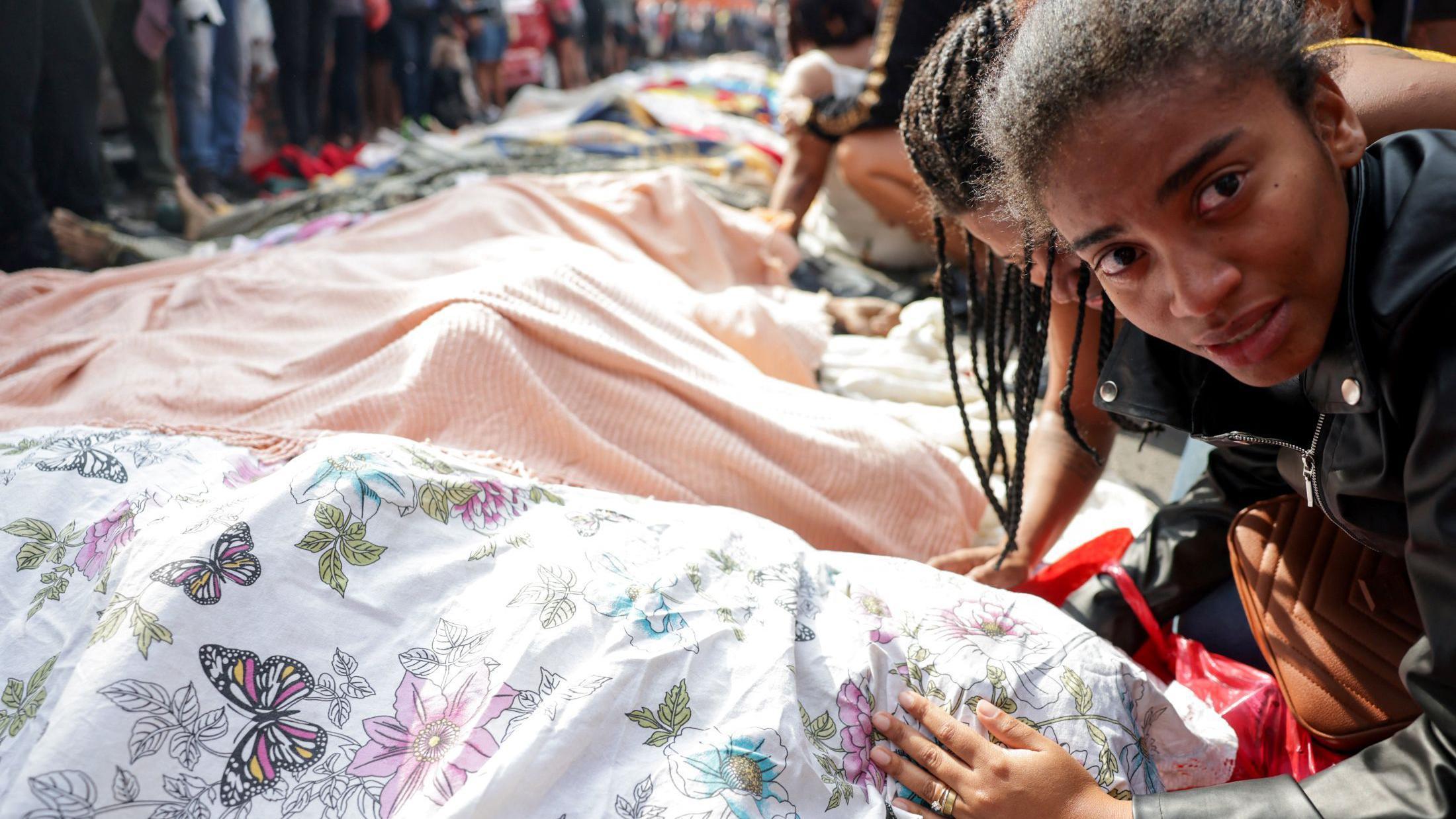 Uma mulher com a mão sobre um corpo coberto com um lençol e enfileirado ao lado de dezenas de outros corpos cobertos. 