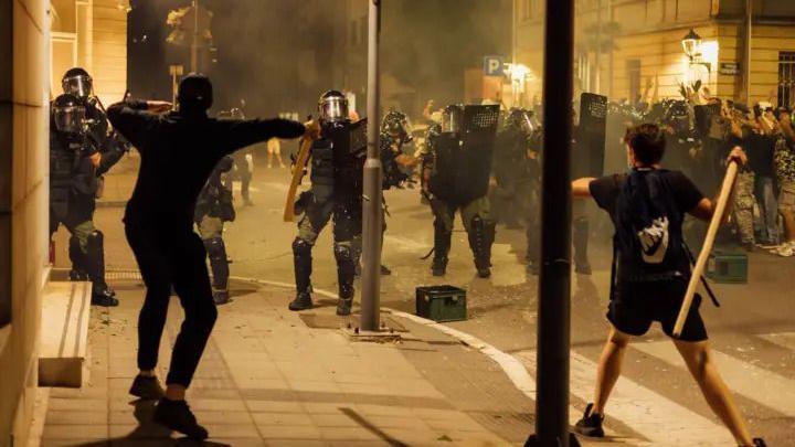 Sa jedne strane policajci sa podignutim štitovima i kacigama na glavi, preko puta njih dva mladića sa drvenim motkama u rukama