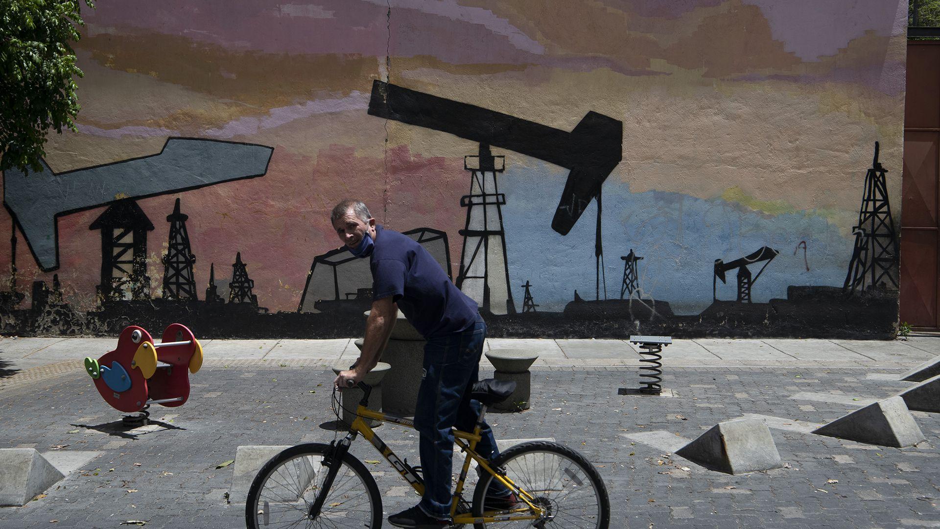 Un hombre en una bicicleta pasa frente a un mural que muestra torres y extracción de petróleo en Caracas en 2020 