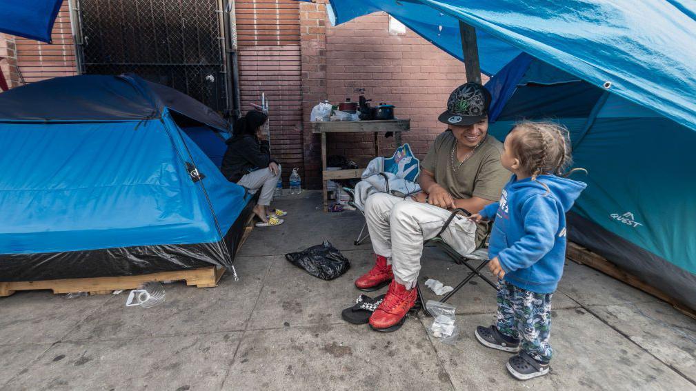 Jhon Valencia juega con Thian, de 2 años, con su madre Katherine González está sentada en una tienda de campaña al fondo, en Towne Avenue en Skid Row, en Los Ángeles, el jueves 25 de abril de 2024. (Myung J. Chun / Los Angeles Times a través de Getty Images)