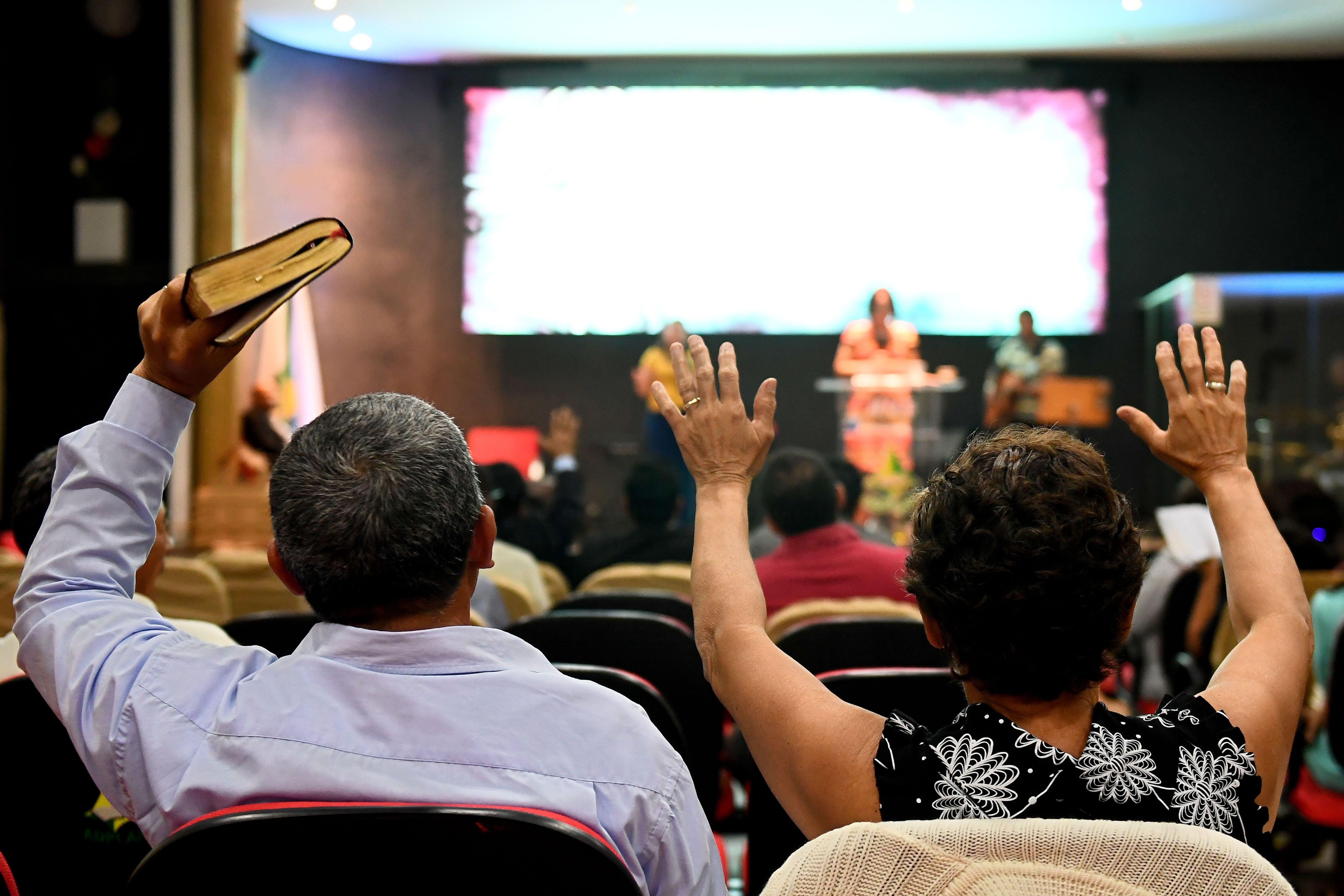 Um homem e uma mulher vistos pelas costas durante culto em igreja evang&eacute;lica. Os dois est&atilde;o sentados de frente para o palco com os bra&ccedil;os erguidos e ele segura uma b&iacute;blia. 