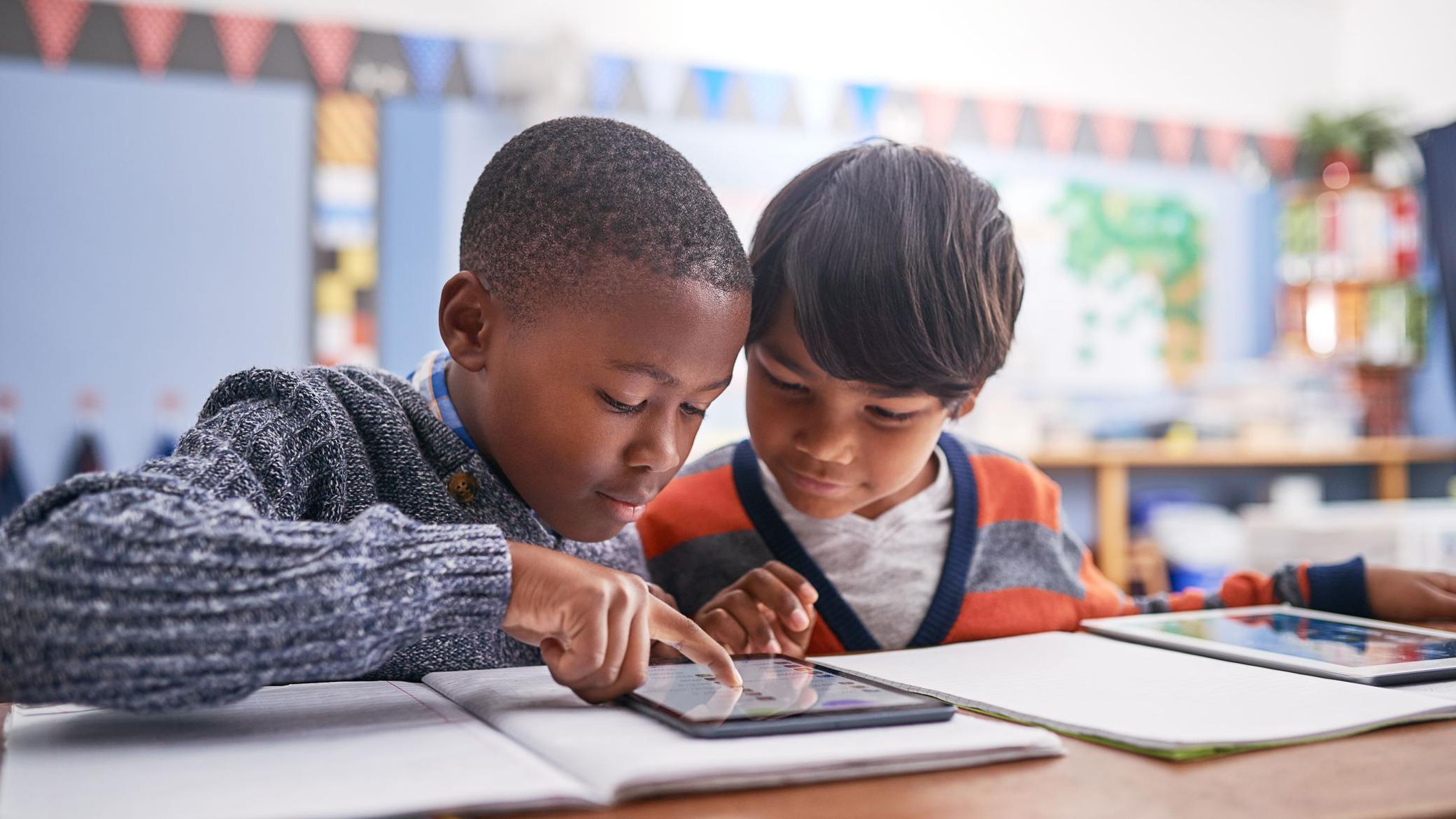 يجلس صبيان جنباً إلى جنب في صف دراسي. ينظر كلاهما إلى جهاز لوحي موضوع فوق دفتر تمارين. يرتدي الصبي على اليسار كنزة رمادية داكنة، بينما يرتدي الصبي على اليمين كنزة مخططة بالبرتقالي والرمادي.