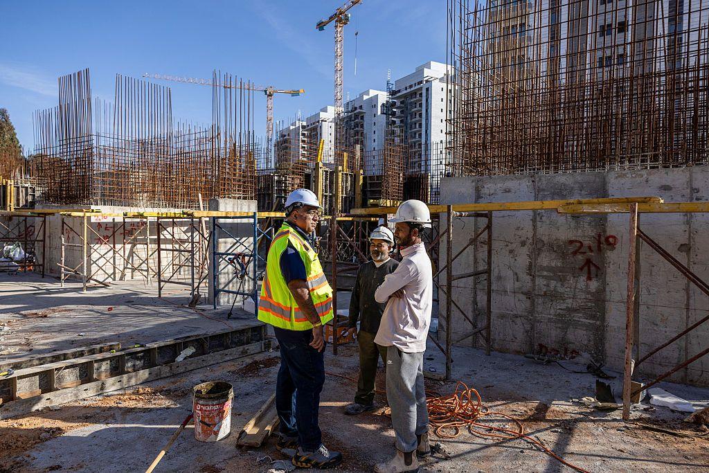 ثلاثة عمال ومهندسين يرتدون خوذات واقية يقفون ويتحدثون داخل موقع بناء، تحيط بهم هياكل حديدية وأعمدة خرسانية قيد الإنشاء، وتظهر في الخلف رافعات ومبانٍ سكنية قيد التطوير، في أجواء عمل بمشروع إنشائي كبير.
