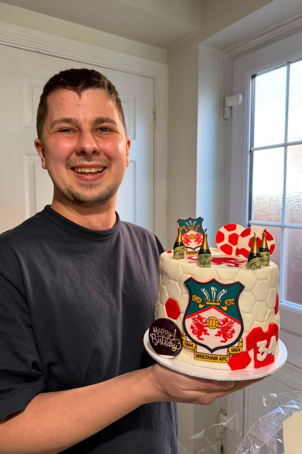 Josh est&aacute; vestindo uma camiseta escura e segurando um bolo de anivers&aacute;rio decorado com a marca do Wrexham FC. Ele est&aacute; sorrindo e olhando para a c&acirc;mera.