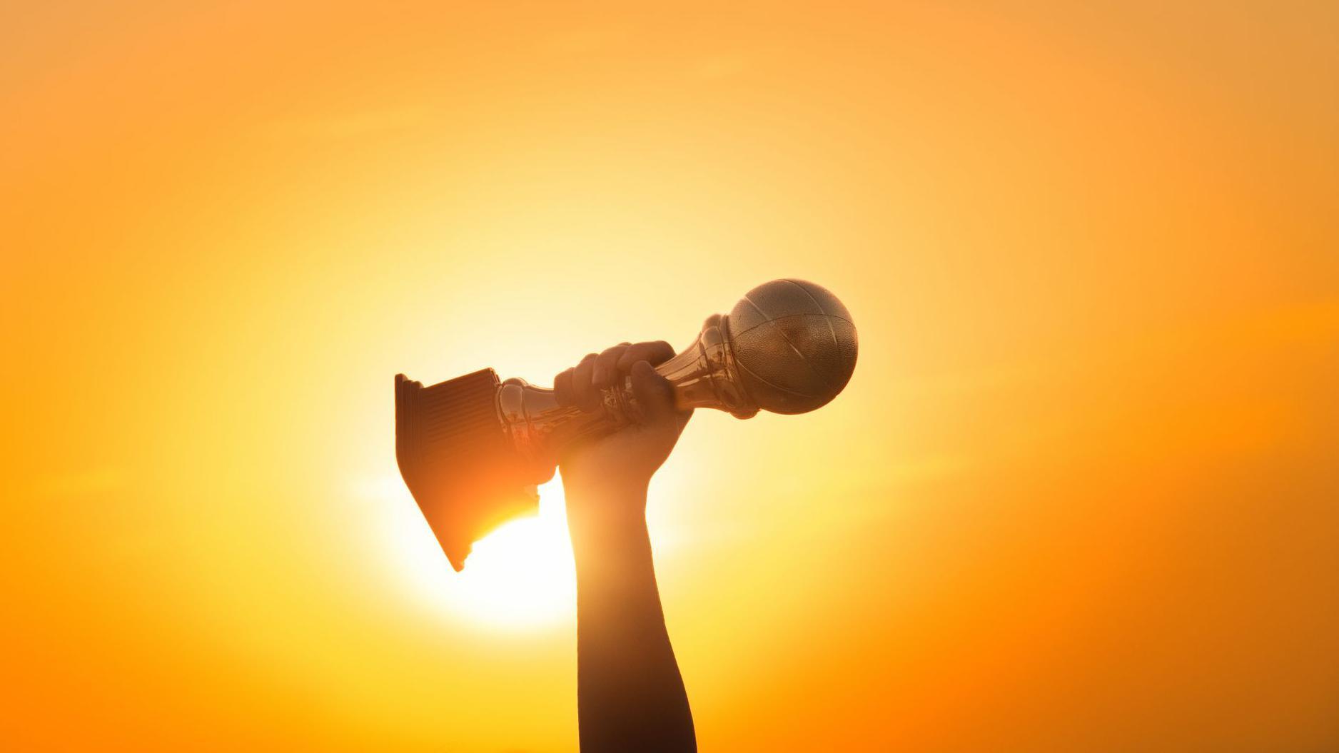 يد ترفع كأس بطولة نحو السماء وخلفها الشمس