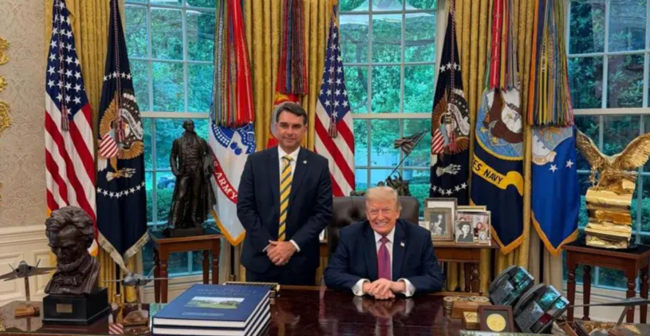 Foto mostrando Donald Trump dentro do sal&atilde;o oval da Casa Branca, com bandeira dos Estados Unidos ao fundo. Fl&aacute;vio est&aacute; em p&eacute; e Trump sentado