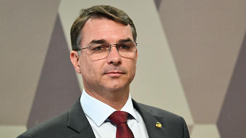 Flavio Bolsonaro &eacute; um homem branco. Ele usa &oacute;culos e est&aacute; com um terno cinza e gravata vermelha.