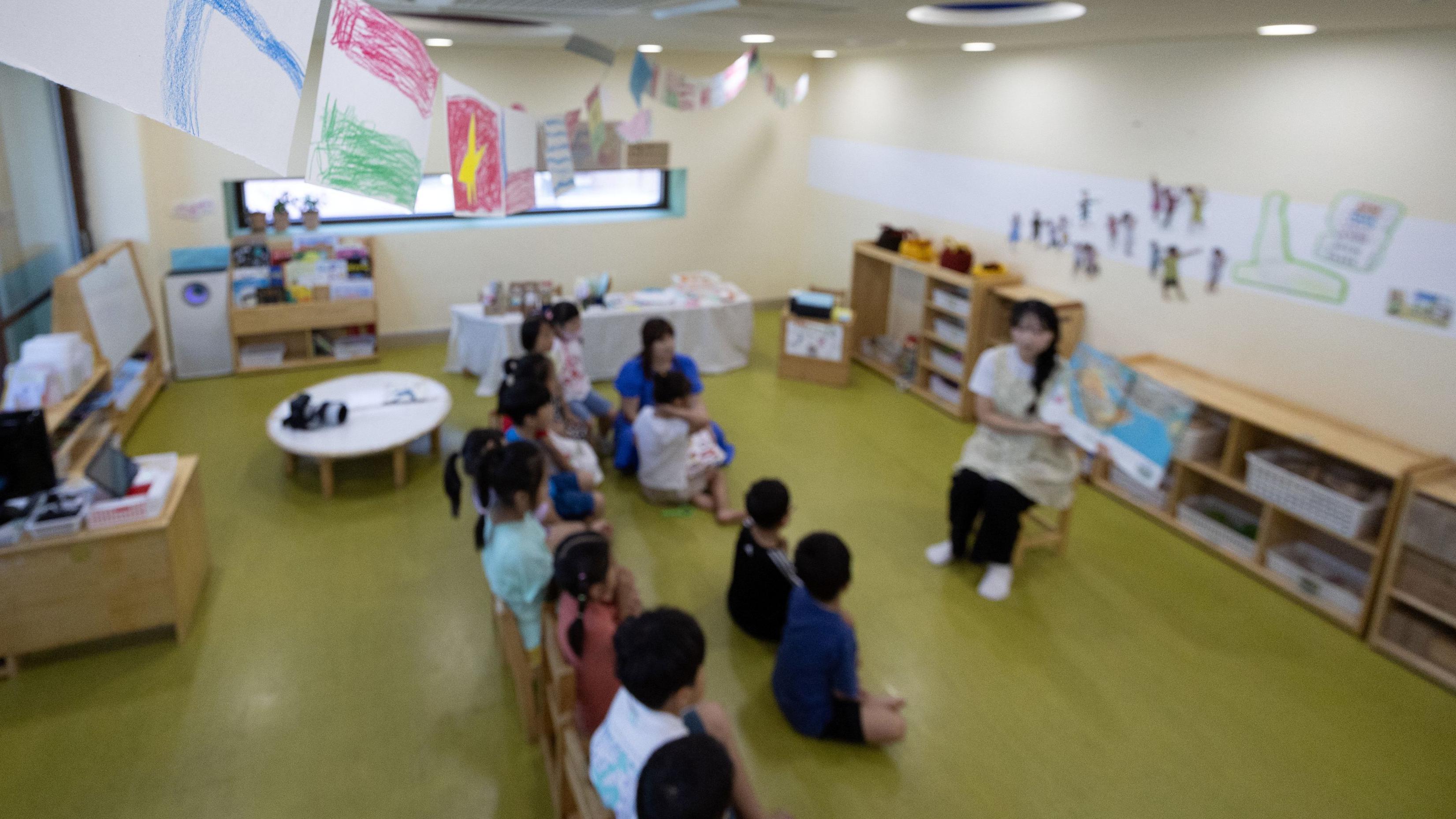 유치원 교실에서 한 교사가 십여명의 아이들 앞에 앉아서 큰 그림 책을 한쪽 팔에 들고 읽어주고 있다 (이 사진은 해당 사건과 관련이 없습니다)