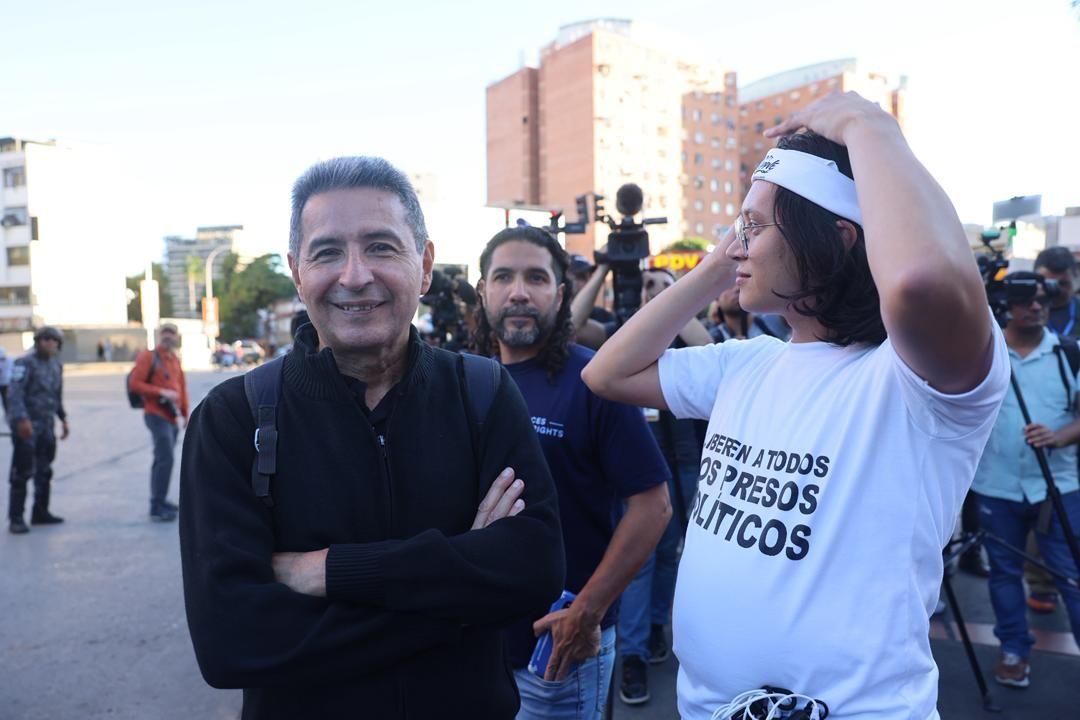Un hombre sonríe a la cámara junto a una persona que lleva una camiseta blanca con el mensaje "Liberen a todos los presos políticos".  