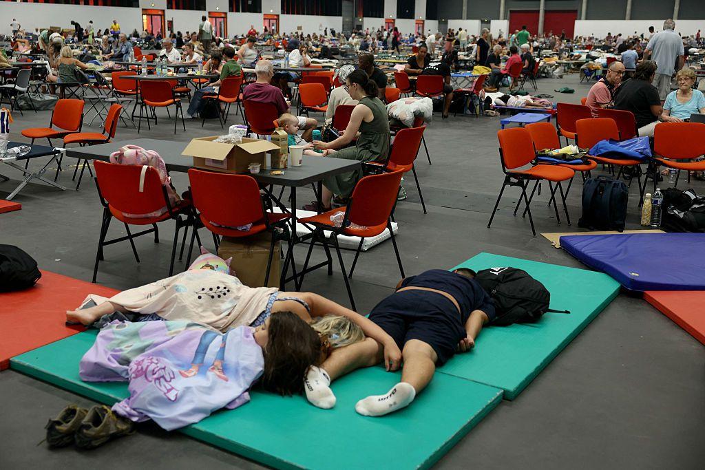 En primer plano, tres personas que parece de la misma familia están echadas sobre colchonetas en el suelo, sin zapatos, tapados con sábanas. Detrás de ellos se ven muchas mesas y sillas donde hay colgadas o puestas mochilas, cajas, enseres varios. Hay infinidad de mesa sy sillas y mucha gente sentada en ellas. 