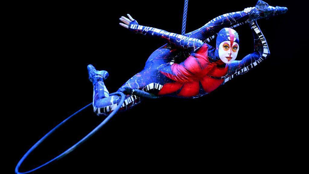 Um artista do Cirque du Soleil, com figurino azul e vermelho e maquiagem completa, se balan&ccedil;a em um trap&eacute;zio enquanto se apresenta com um bambol&ecirc;