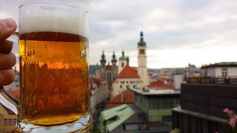 Mão segurando uma caneca com cerveja. No fundo, cidade de Praga