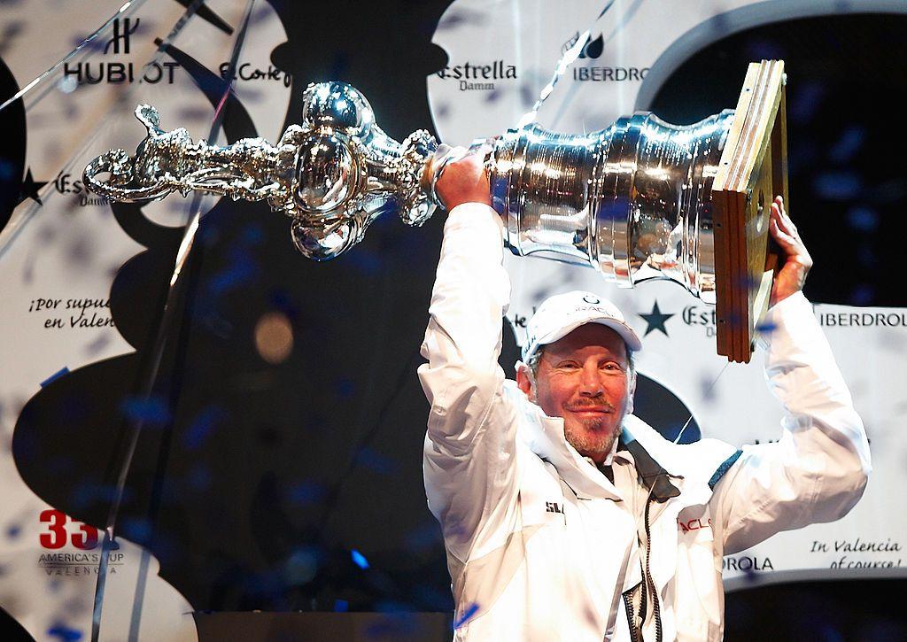 Larry Ellison, vestido con gorra y ropa deportiva blanca, levanta un gran trofeo plateado con ambas manos.