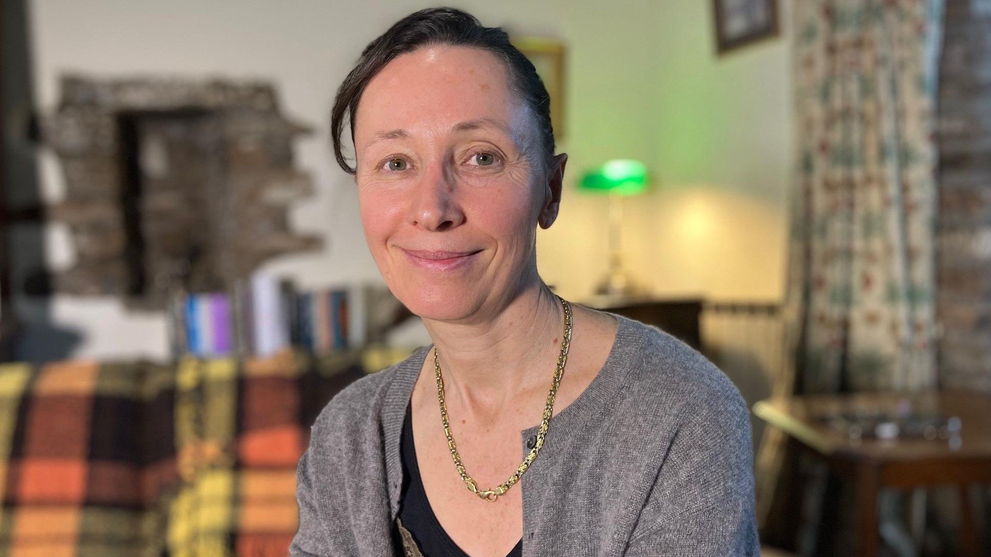 Dr Hard sitting on her sofa wearing a grey cardigan and gold necklace