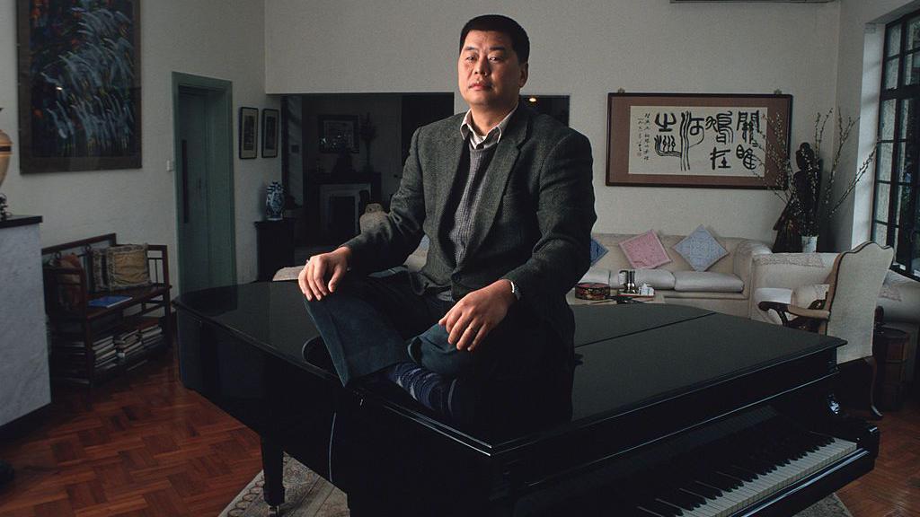 Jimmy Lai, wearing a suit jacket and vest, sits above a grand piano at his home from a picture taken in 1993