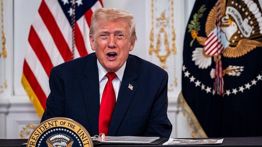 Donald Trump sentado en un escritorio, con las banderas de Estados Unidos y de la Presidencia de Estados Unidos en el fondo