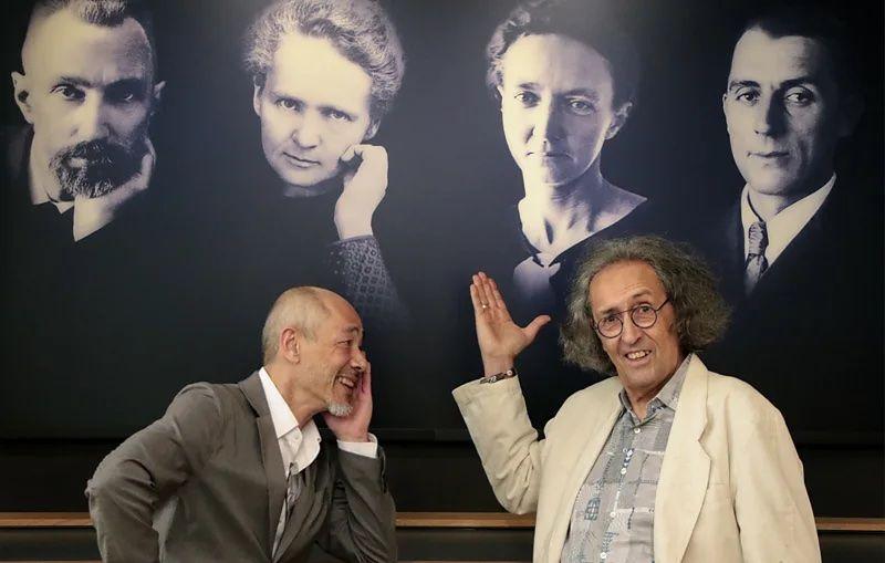 Dois homens estão em frente a um quadro de fotos que mostra 4 pessoas da família Curie