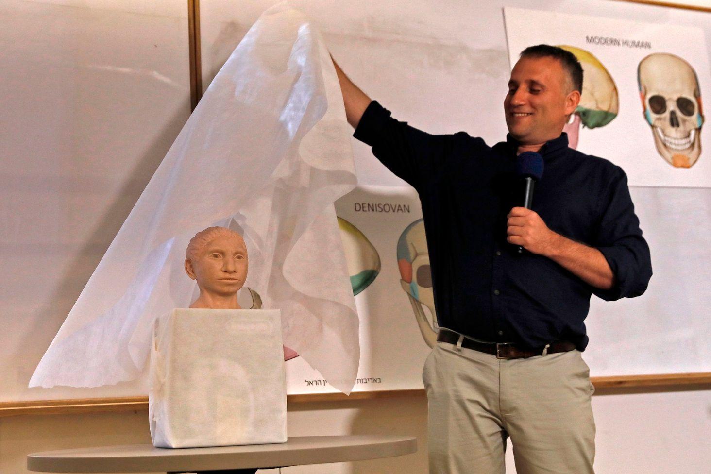 El profesor Liran Carmel, con una camisa negra y un pantalón beige, levanta una cubierta blanca y deja al descubierto el modelo de un rostro beige