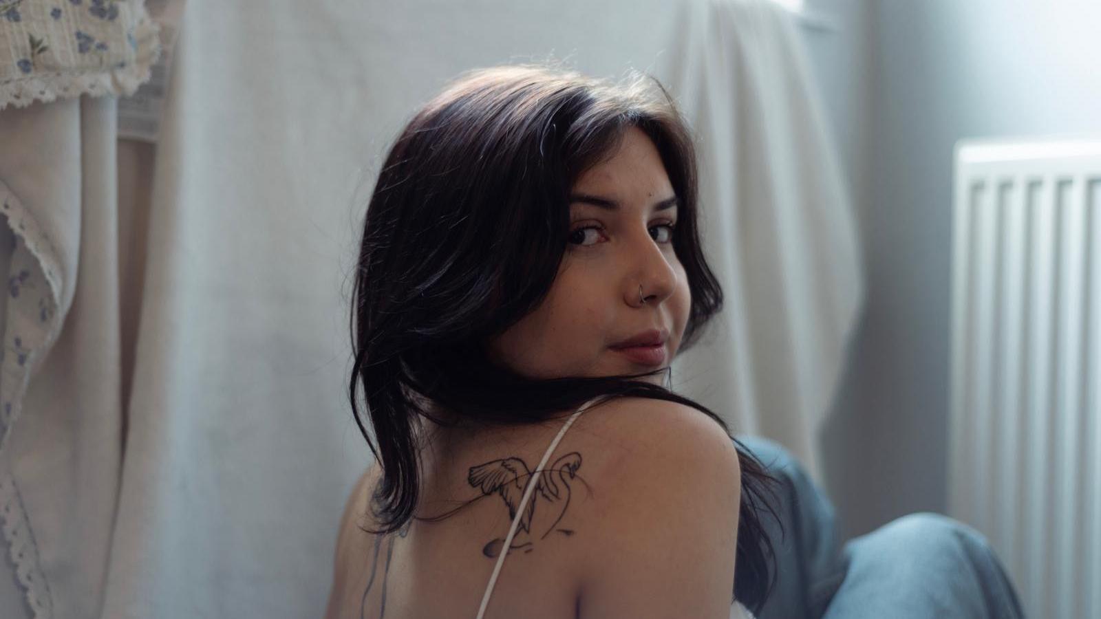 A young woman with dark hair looks back over her shoulder at the camera. She has a tattoo of a sw