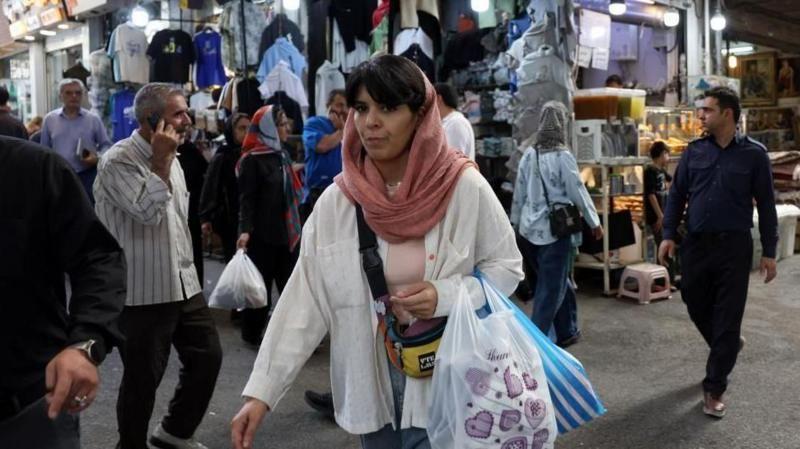 Mujer lleva una bolsa con motivos violeta en medio de un mercado donde se destaca una tienda con 