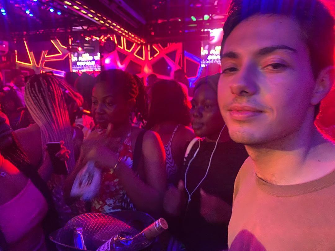 Carlos se encuentra en una discoteca abarrotada, vistiendo una camisa de color claro bajo una intensa iluminación de neón rosa, morada y azul. Está a la derecha de la foto; a su lado hay varias mujeres y una cubitera metálica con botellas.