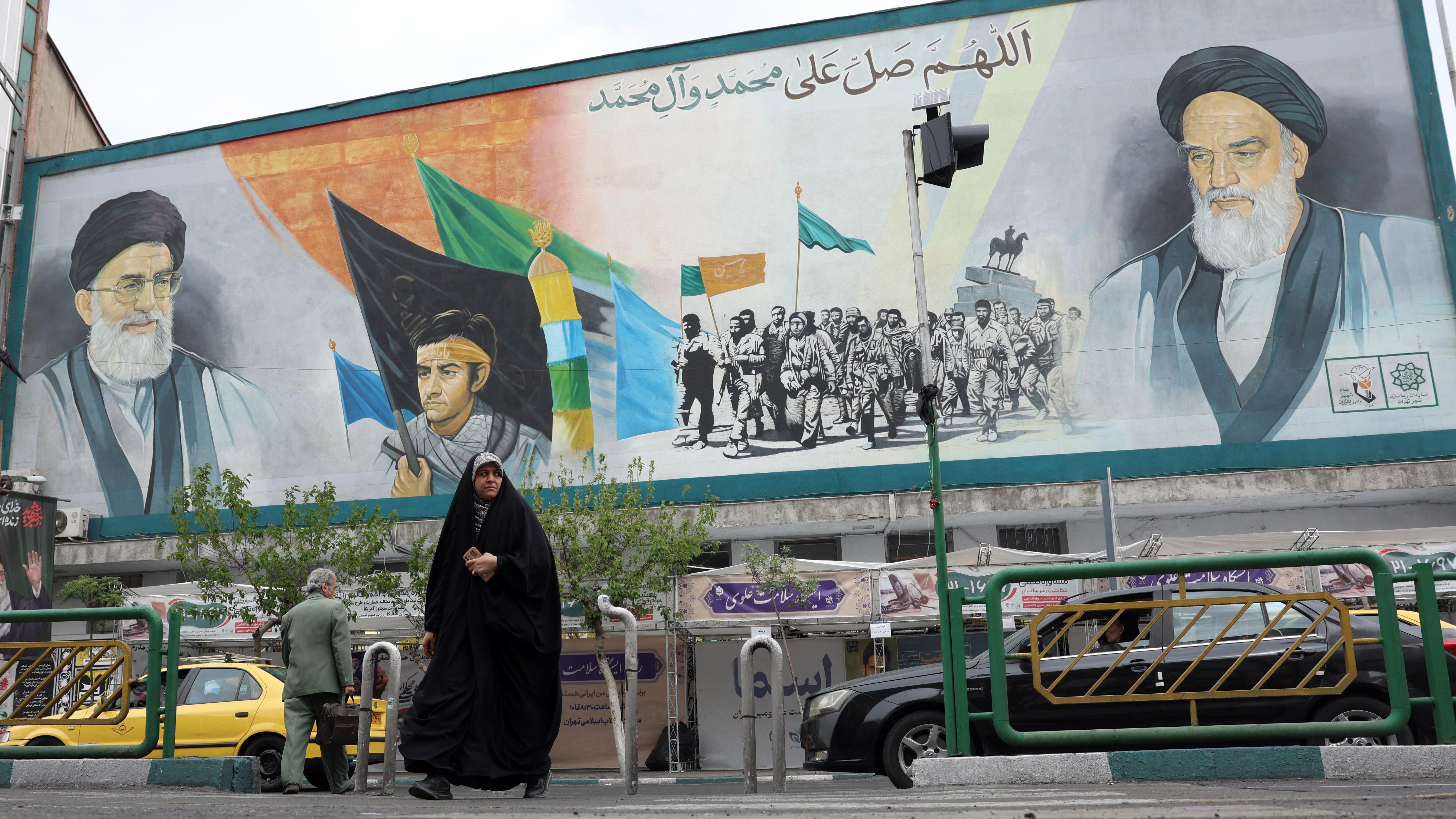 伊朗德黑蘭街頭一位身穿全身罩袍但露臉的女士在一幅帶有已故最高精神領袖哈梅內伊與霍梅尼的畫像前走過（12/4/2026）