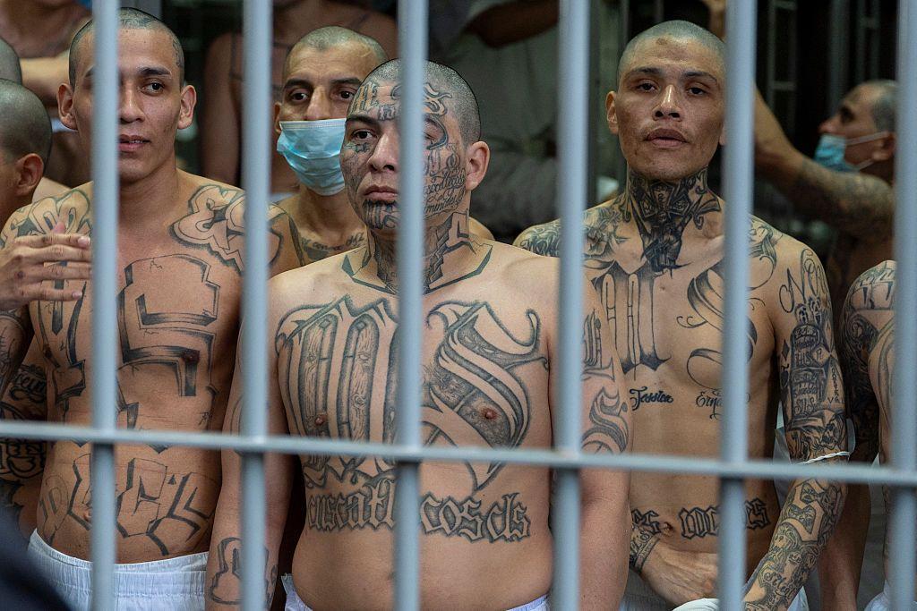 Detrás de las rejas de una celda en la cárcel salvadoreña del Cecot, se puede ver a tres hombres en primer plano sin camisas, con el pecho y los brazos tatuados. El del centro y el de la izquierda tienen tatuadas las siglas MS, usadas por la Mara Salvatrucha.