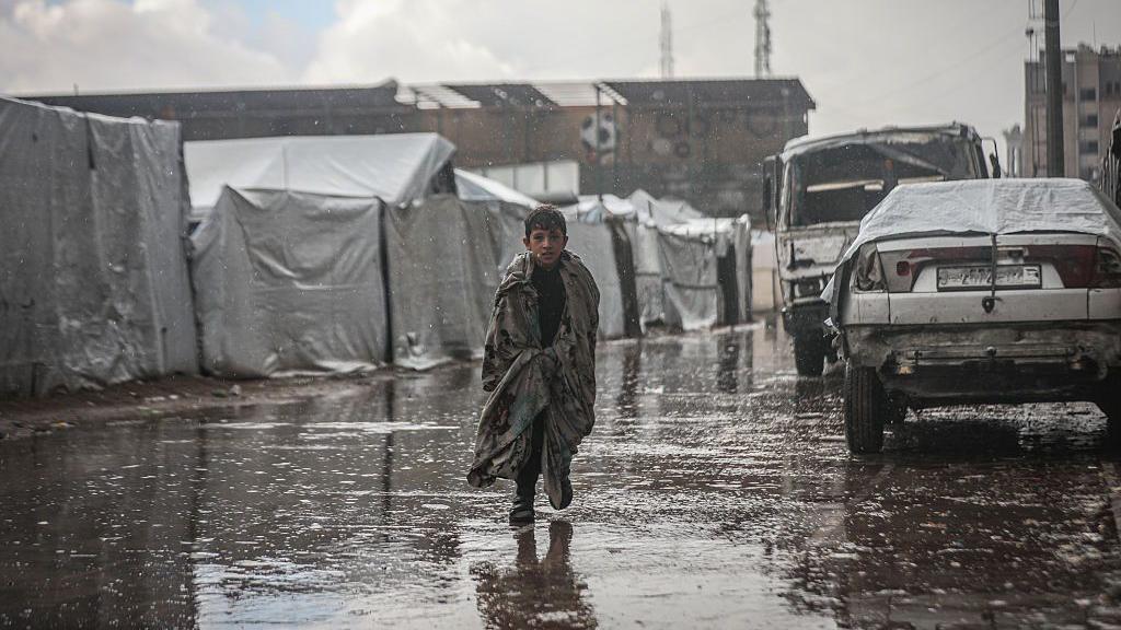 طفل فلسطيني يركض تحت المطر في غزة بينما تظهر خيام النازحين من خلفه وقد غمرتها المياه