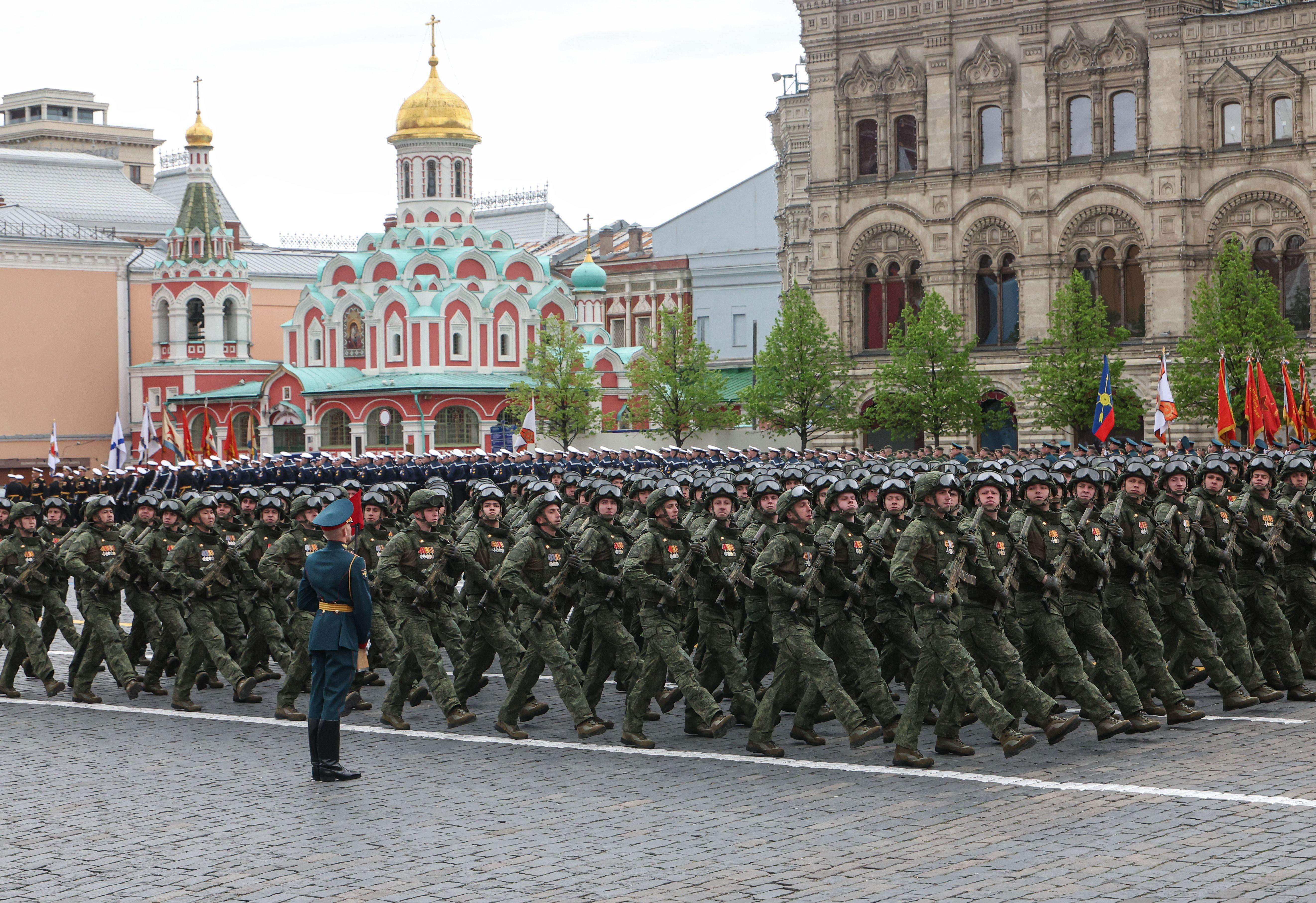 Unos militares rusos desfilan en filas vestidos con uniformes de camuflaje verde oscuro, cascos con gafas protectoras colocadas encima y empuñando armas. Detrás de ellos se divisa la catedral de Kazán, pintada en tonos verde azulado, rojo y blanco, y coronada por una cúpula dorada.