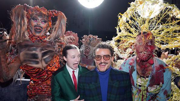 A imagem mostra um grupo de pessoas posando para fotos em um evento, cercadas por performers caracterizados como criaturas infectadas do universo de The Last of Us. &Agrave; esquerda, h&aacute; um ator fantasiado com um figurino grotesco, composto por grandes placas de fungo e tecidos deformados que cobrem todo o corpo. &Agrave; direita, outro performer veste uma fantasia complexa, com uma grande estrutura dorsal que se assemelha a ramifica&ccedil;&otilde;es de fungo crescendo para fora da cabe&ccedil;a e das costas, criando um visual expansivo e detalhado. As fantasias s&atilde;o ricas em texturas, com cores que variam entre tons avermelhados, amarelados e marrons, lembrando carne infectada e crescimento f&uacute;ngico. Os trajes chamativos contrastam com as roupas formais das pessoas no centro da imagem, refor&ccedil;ando o clima tem&aacute;tico da pr&eacute;?estreia da s&eacute;rie.