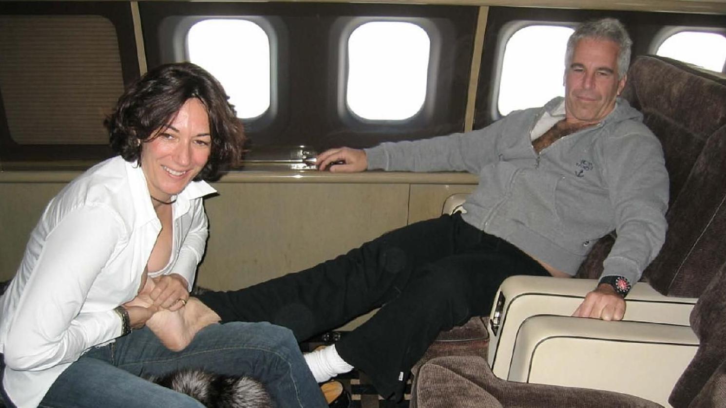 Ghislaine Maxwell and Jeffrey Epstein on board a plane. Epstein is lounging in a seat with his legs stretched out. Maxwell is leaning forwards in a seat opposite him. She is smiling, while he has his mouth closed. Maxwell is wearing a white top and blue jeans. Epstein is wearing a grey hooded sweatshirt and black trousers.