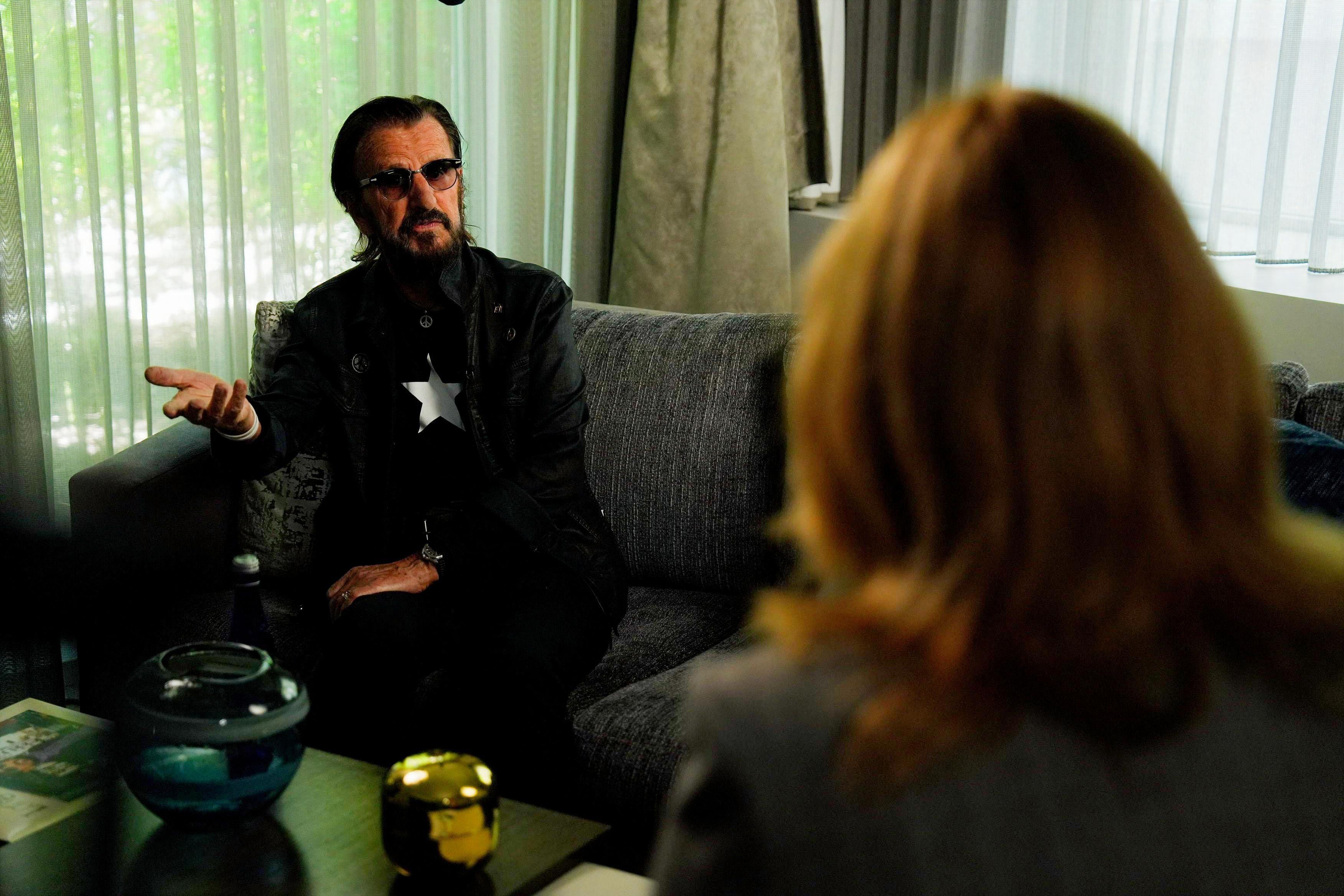 Ringo Star u prvom planu sedi na kauču i razgovara sa ženom okrenutom leđima ka kameri
